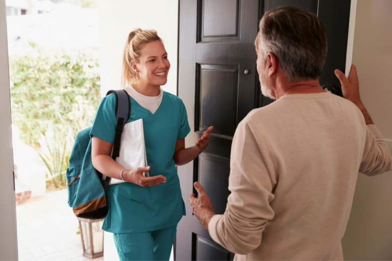 A nurse in teal scrubs is greeted by a patient at their home.