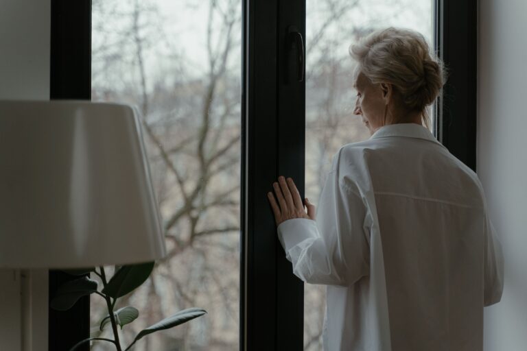 An older adult looks outside of a window as the weather starts to shift towards Fall